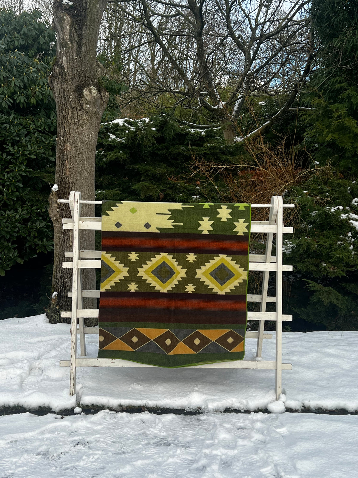 Alpaca deken Otavalo met geometrisch patroon in groen, crème, geel en roestbruin, op een wit rek in de sneeuw